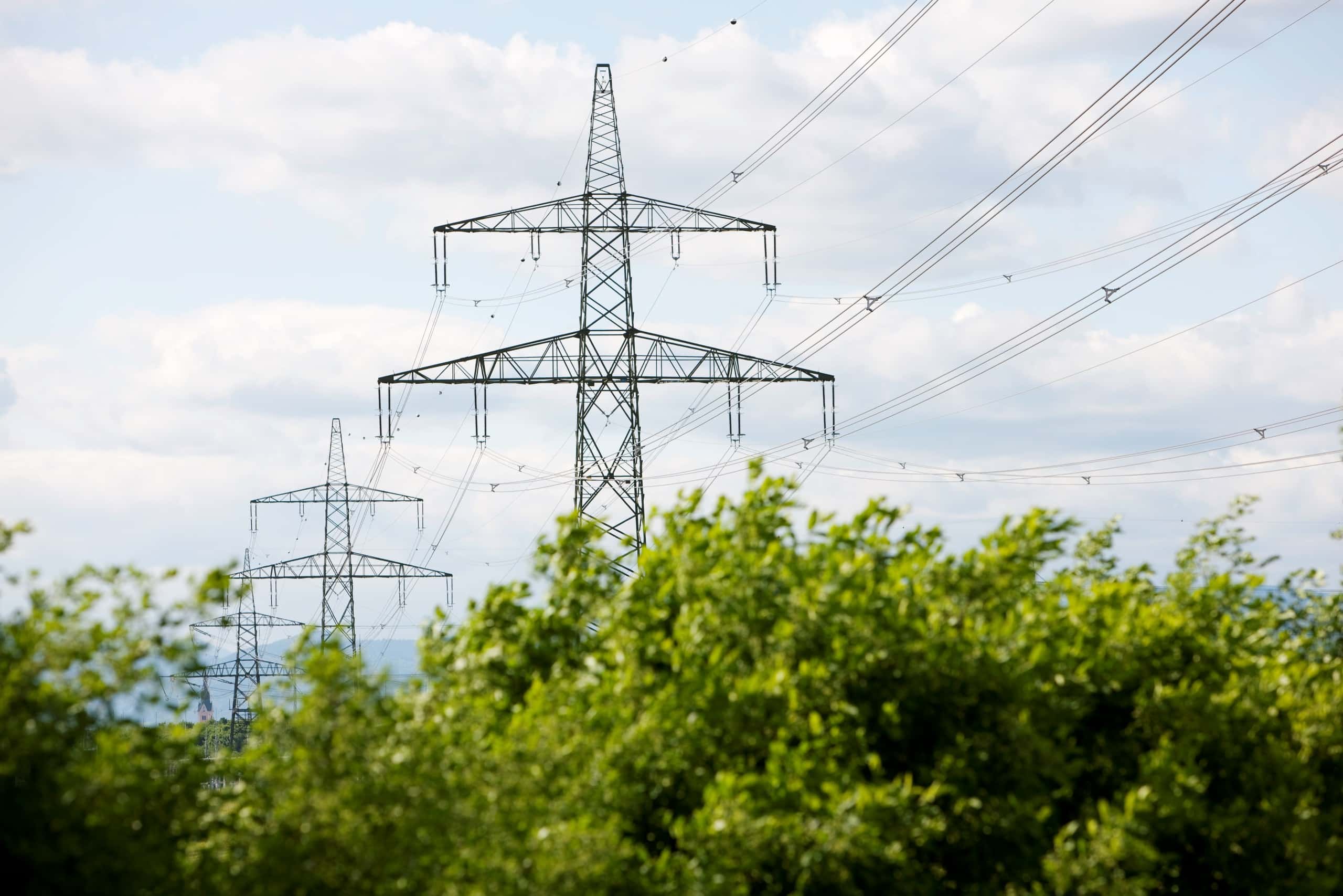Zwei große Hochspannungsmasten in grüner Landschaft mit sichtbaren Stromleitungen unter teils bewölktem Himmel
